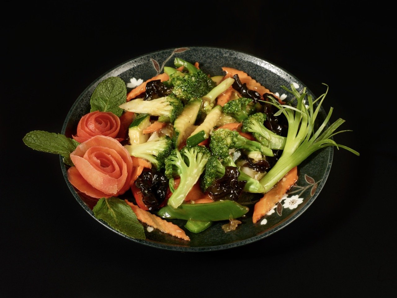 Salade de légumes frais avec des carottes, brocolis, céleri, feuilles de menta, et fleurs de carotte en forme de roses, dans un bol noir avec des motifs floraux.
