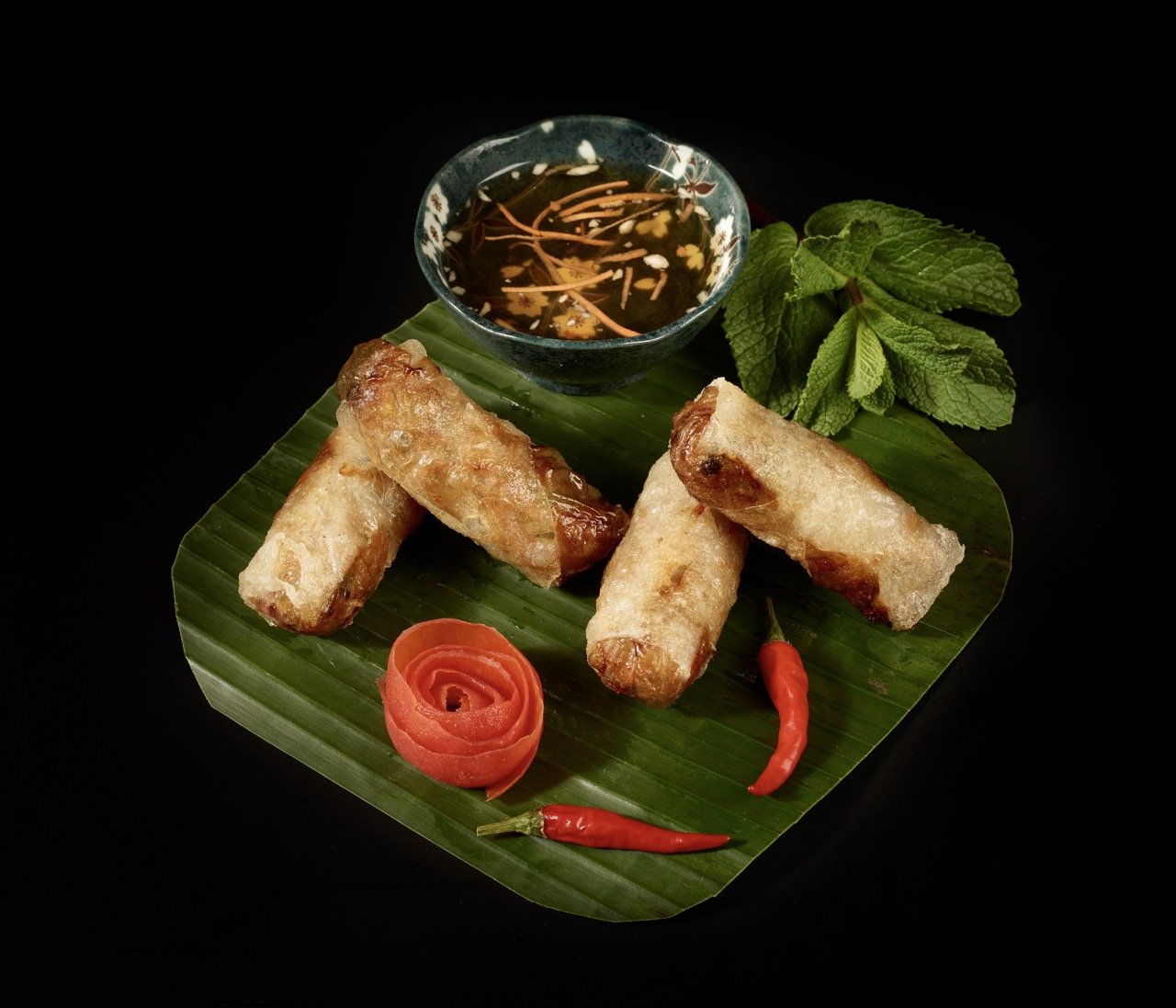 Rouleaux de printemps frits servis avec une sauce à tremper, de la coriandre fraîche, une rose en tomate, deux petits piments rouges, et une branche de menthe, le tout présenté sur une feuille de bananier noire.