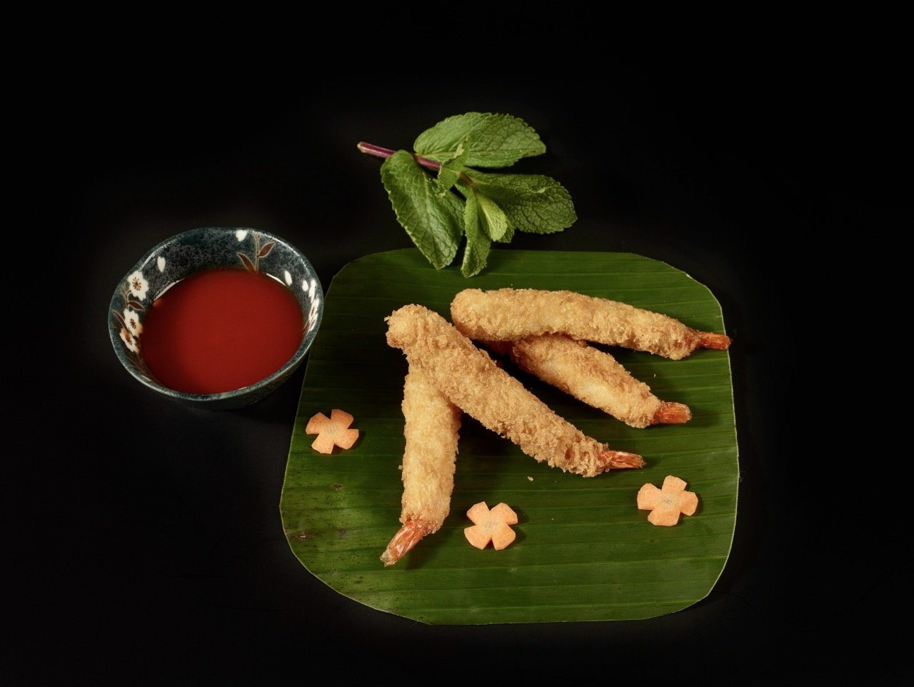 Gambas tempura dorées servies sur une feuille de bananier, accompagnées d'une sauce rouge dans un bol décoratif, avec des feuilles de menthe et des fleurs en décoration.