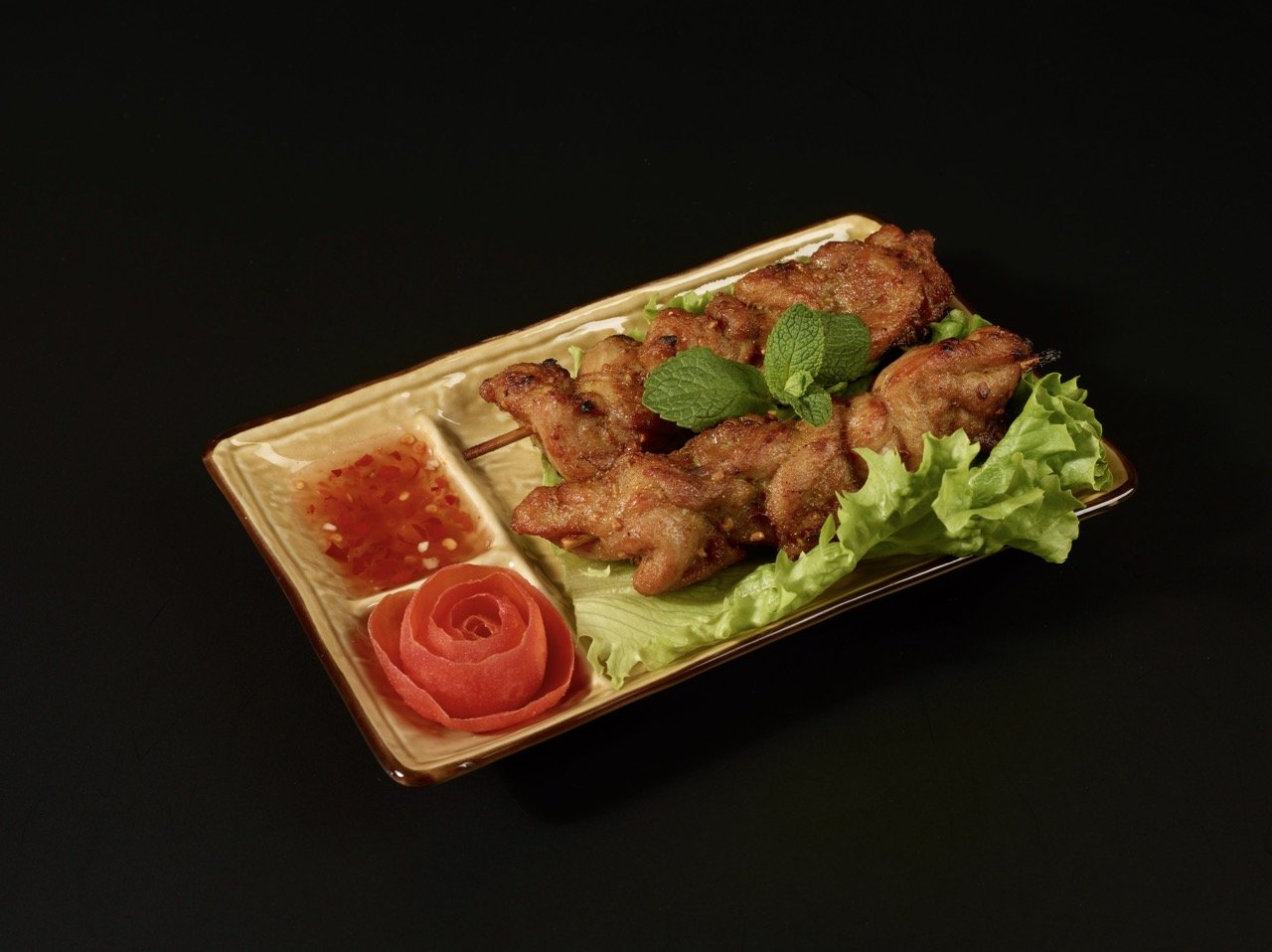 Brochette de viande servie avec du riz thaï à la sauce piquante, une feuille de laitue, une rose faite avec une tranche de tomate, et une branche de menthe sur une assiette rectangulaire beige, sur fond noir.