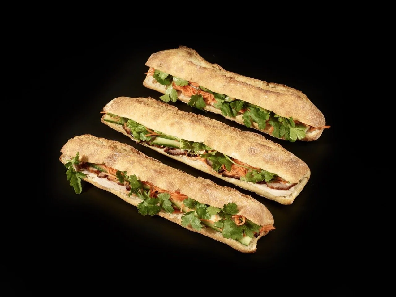 Trois sandwiches baguette avec viande, carottes, coriandre et légumes frais sur un fond noir.