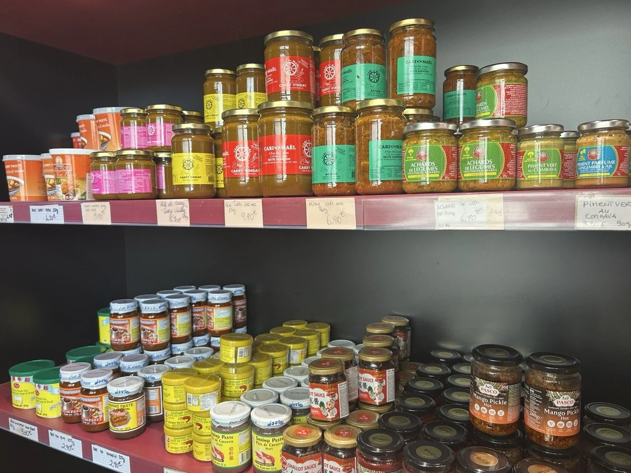 Rayonnage de pots de condiments et sauces, notamment des pâtes de crevettes, mangues pickles, sauces piquantes, et autres produits en conserve de différentes couleurs et tailles.