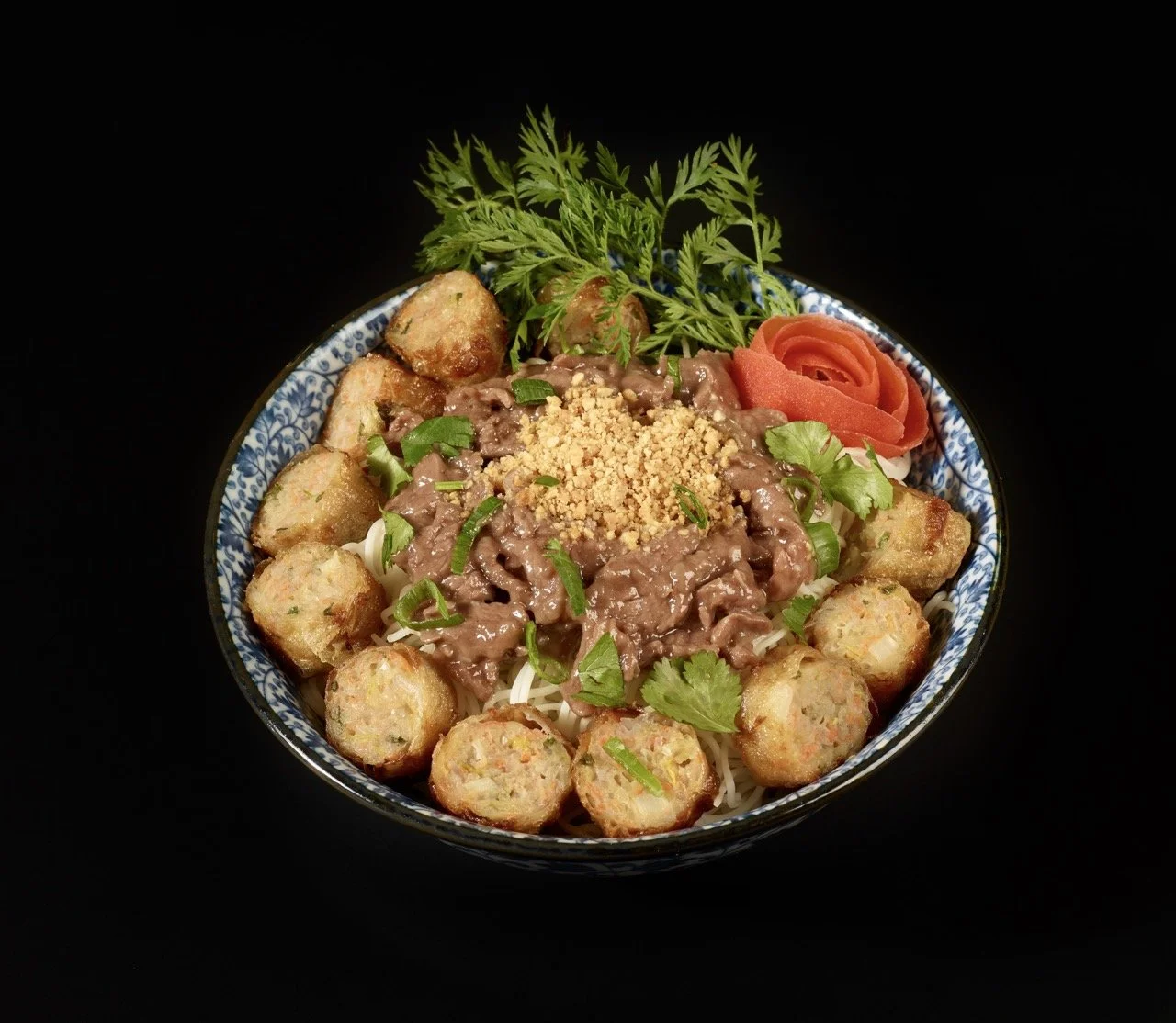 Bol de nourriture asiatique avec des boulettes frites, de la viande en sauce, des herbes fraîches, une tranche de tomate enroulée, et des cacahuètes moulues, dans un bol bleu et blanc.