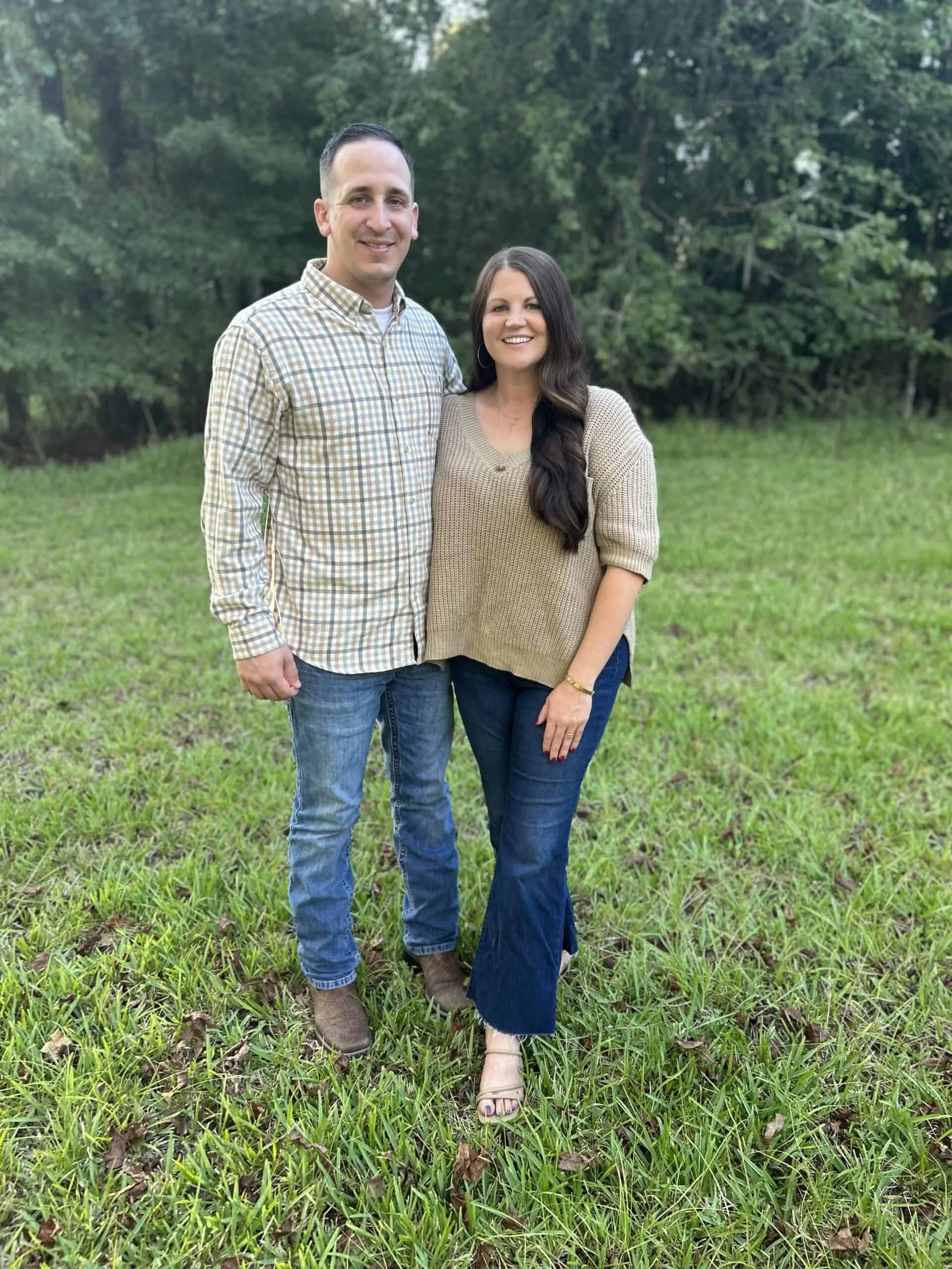 A smiling man and woman standing closely together outdoors on a grassy area with trees in the background. The man is wearing a plaid shirt and jeans, and the woman is wearing a beige sweater and dark jeans.