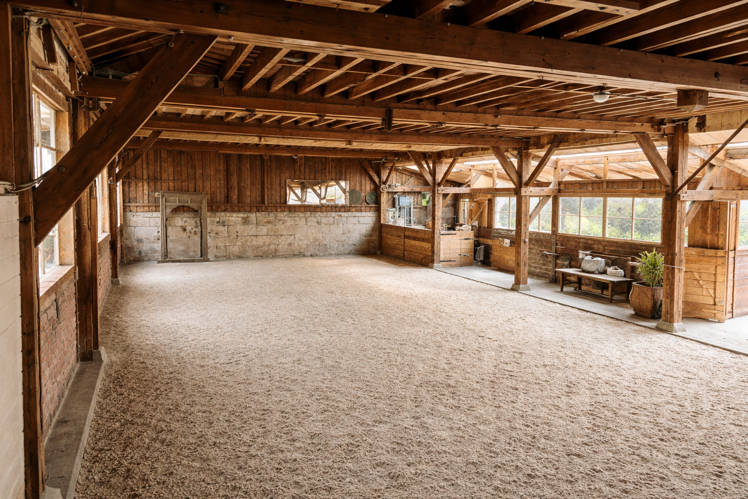 Intérieur d'une grande grange en bois avec sol en tapis beige, murs en pierre et grosses poutres apparentes, fenêtres offrant une vue sur la nature.
