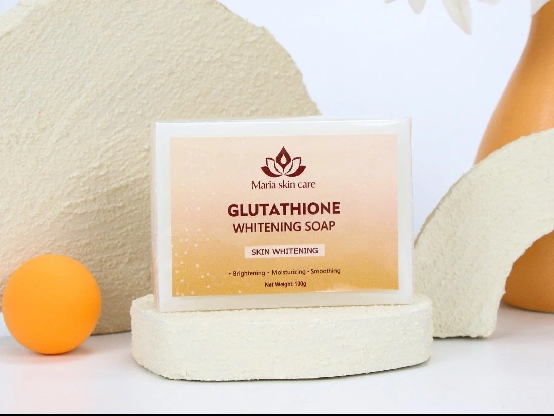 A bar of Maria Skin Care Glutathione Whitening Soap on a white round platform with geometric decor in the background, including a yellow ball and beige textured objects.