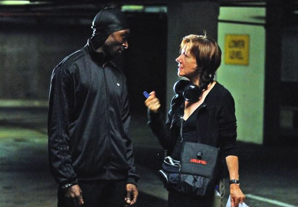 A man and woman in a conversation in what appears to be an indoor parking lot or garage. The man is wearing a black jacket and a black beanie, while the woman has short brown hair and is holding a set of headphones around her neck and a small bag.