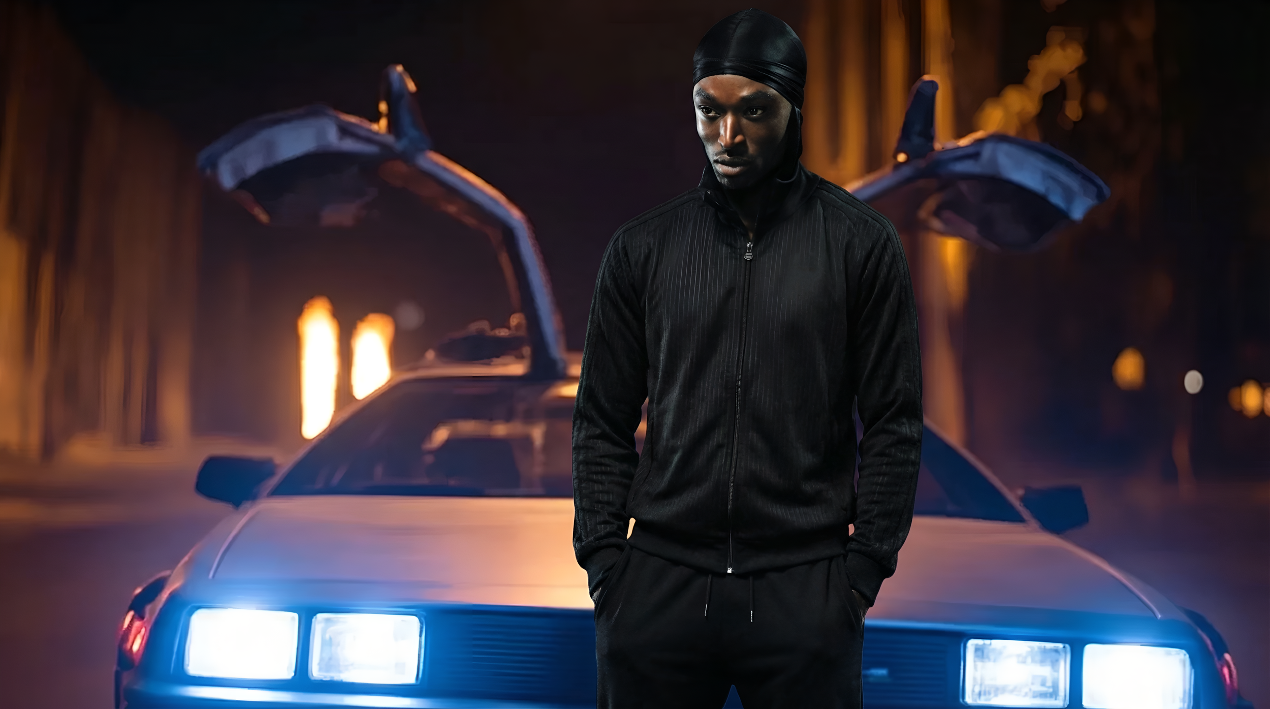 A young man in a black zip-up jacket and black head covering standing in front of a sports car with open gullwing doors, illuminated by streetlights and city lights at night.