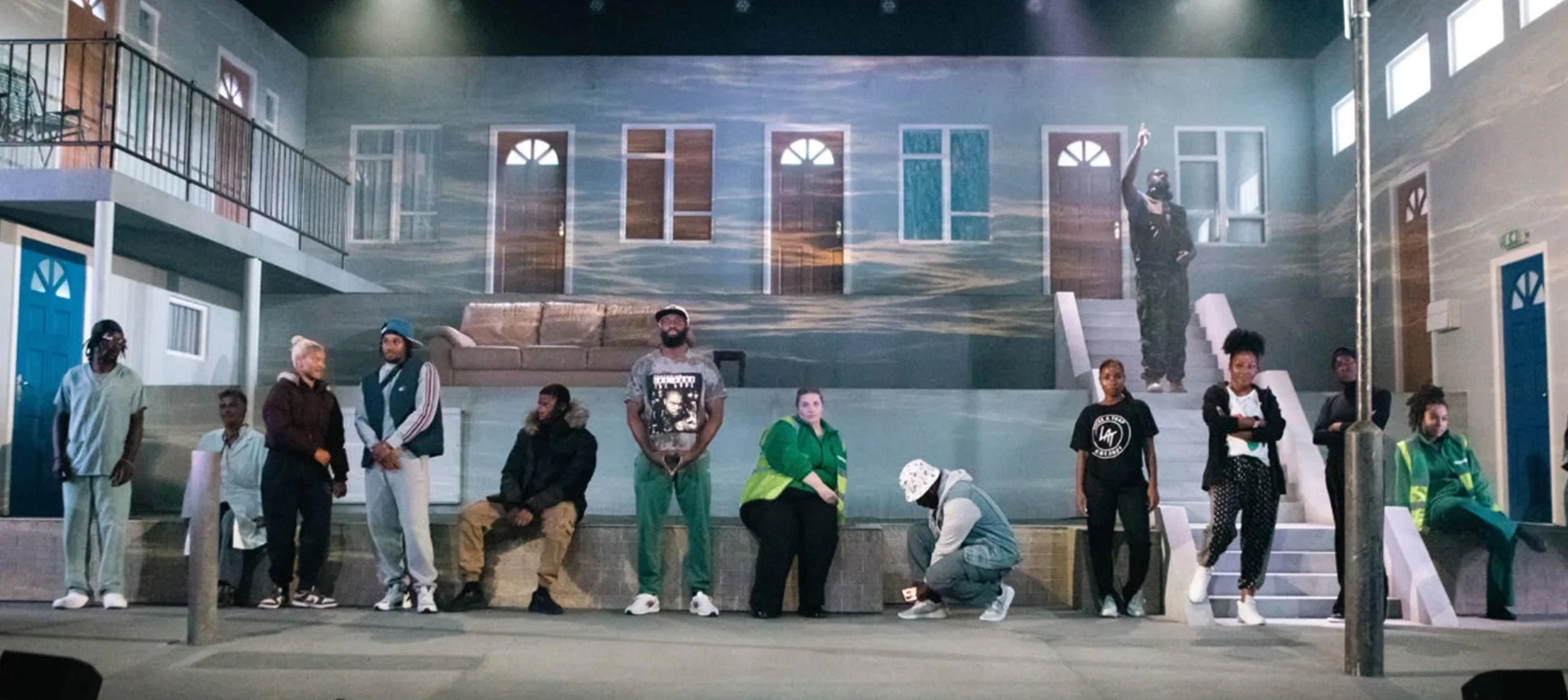 A group of diverse people standing and sitting in front of a mural depicting a house with multiple windows, a staircase, and a brown couch, in an indoor setting with artificial lighting. 4 walls play Derby.