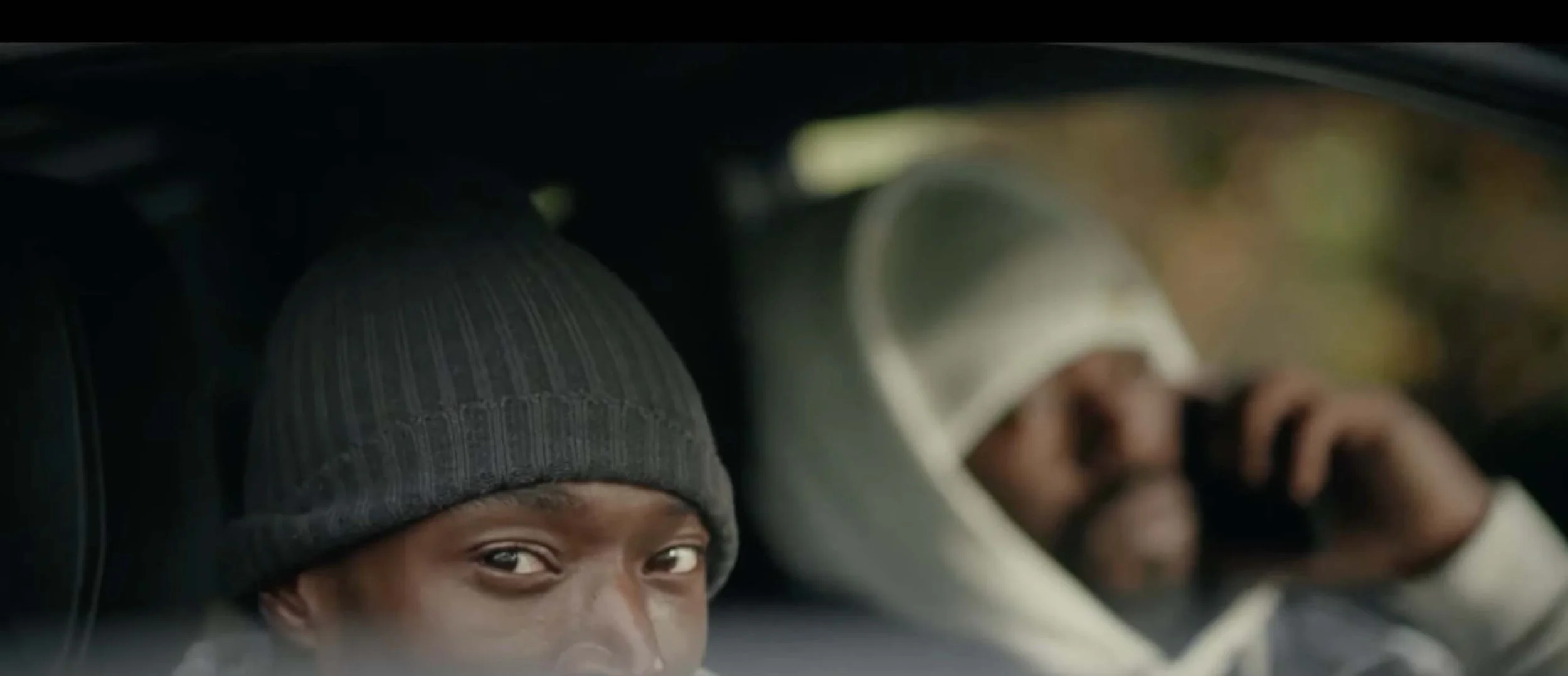 Close-up of two people in a car, one wearing a gray knit beanie and smiling, the other in a white hoodie, with blurred window view.