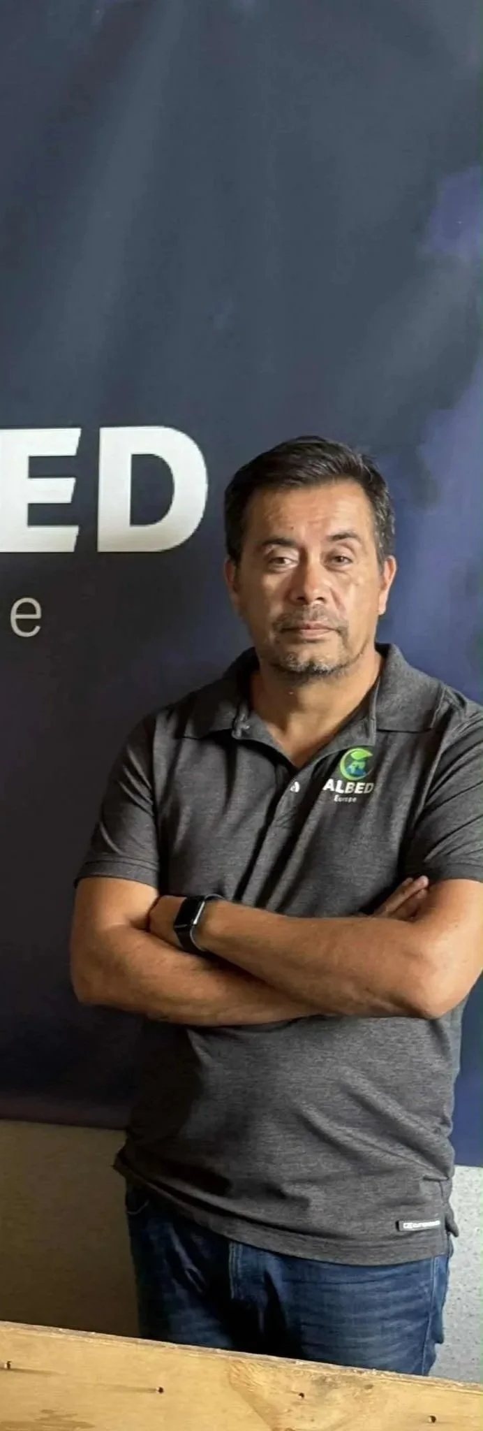 A man with dark hair and a beard, wearing a gray polo shirt with a logo, stands with arms crossed in front of a dark promotional background with partial text.