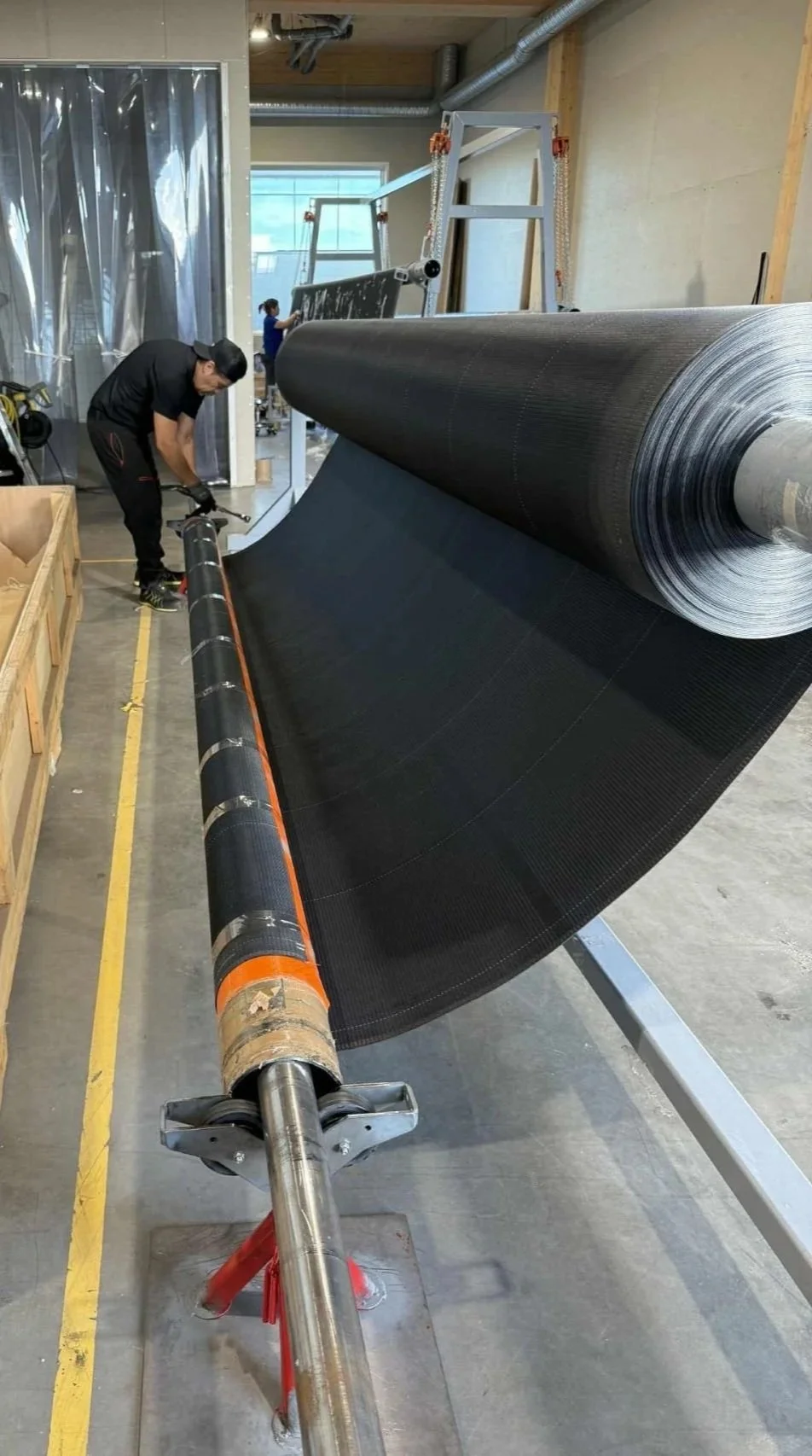 Workers installing a large black flexible material, possibly a roofing or insulation membrane, on a roll in an industrial setting.