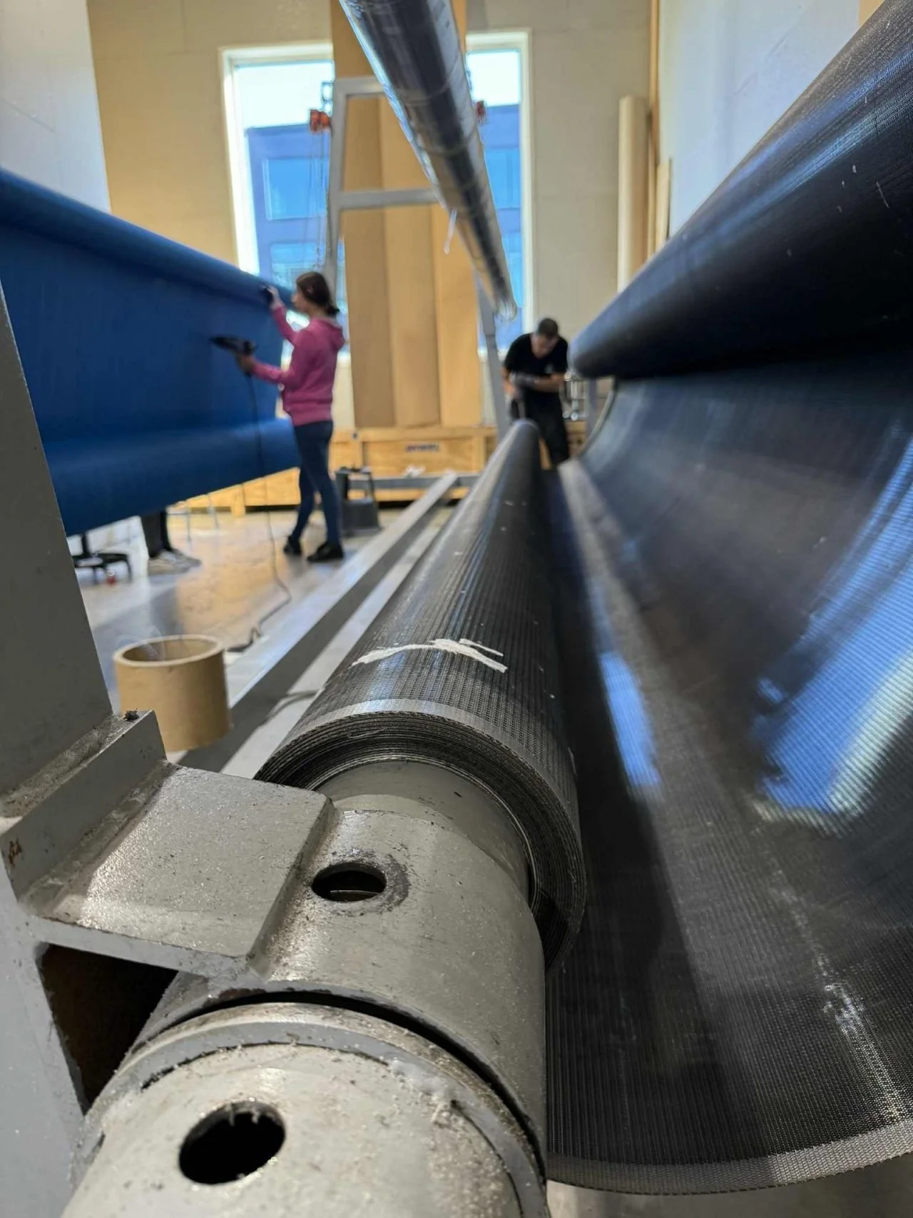 Close-up of large rolled-up black material, with people working in the background in a workshop or manufacturing setting.