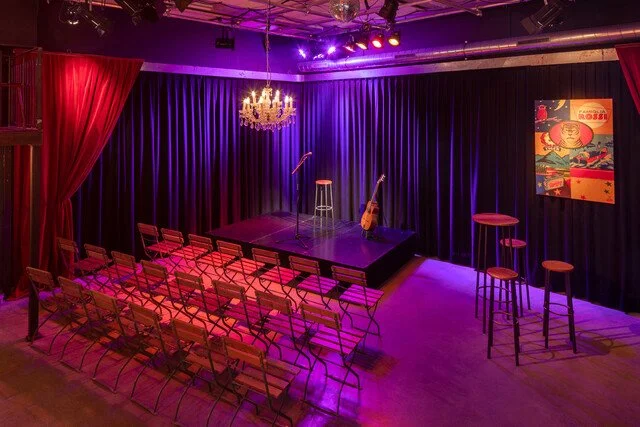 Bühne mit Mikrofonen, Gitarre und Stühlen, im Hintergrund schwarze Vorhänge, bunte Beleuchtung und Dekoration