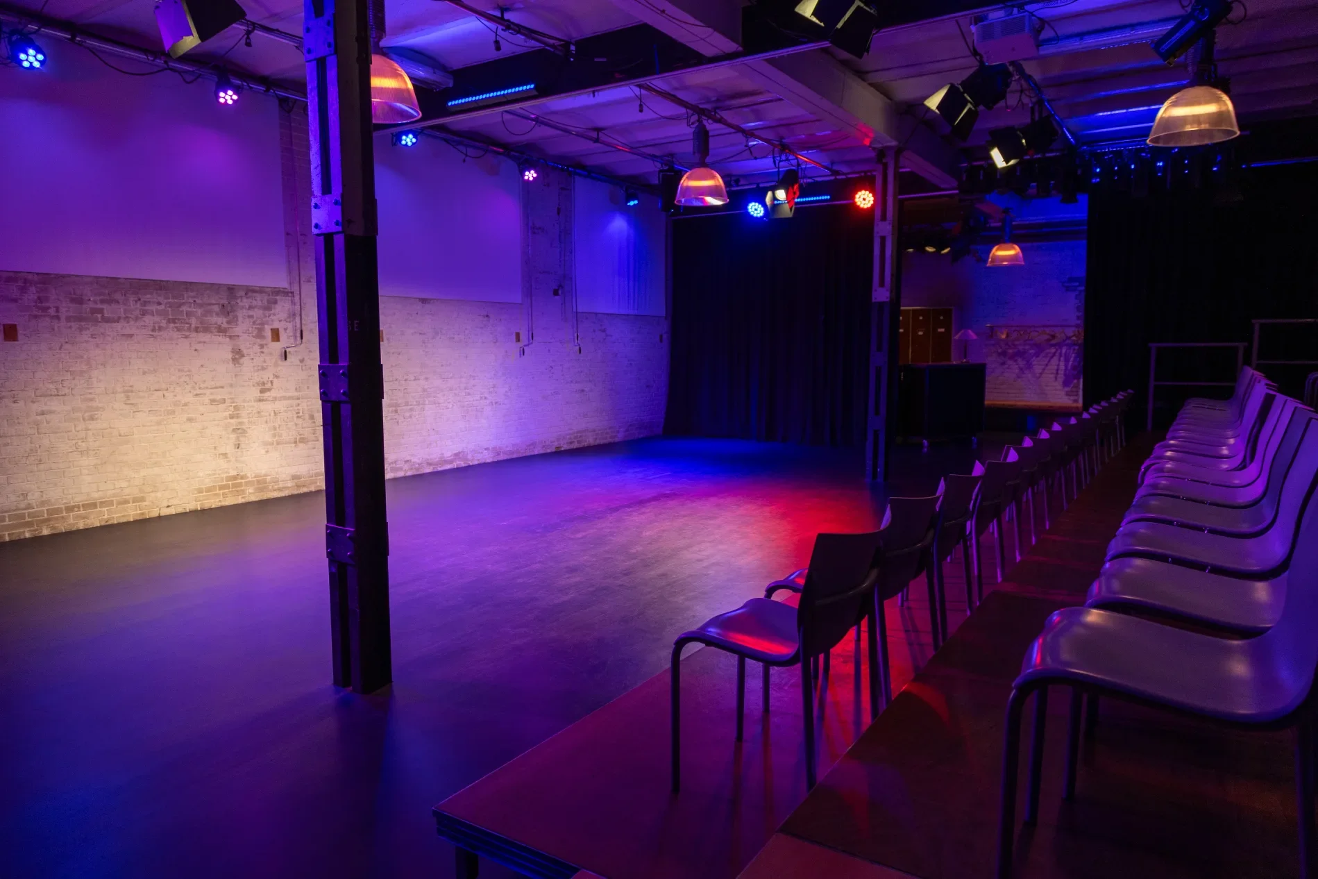 Leerer Veranstaltungsraum mit Stühlen in einer Reihe, Bühne, bunte Bühnenbeleuchtung und schwarzer Vorhang.