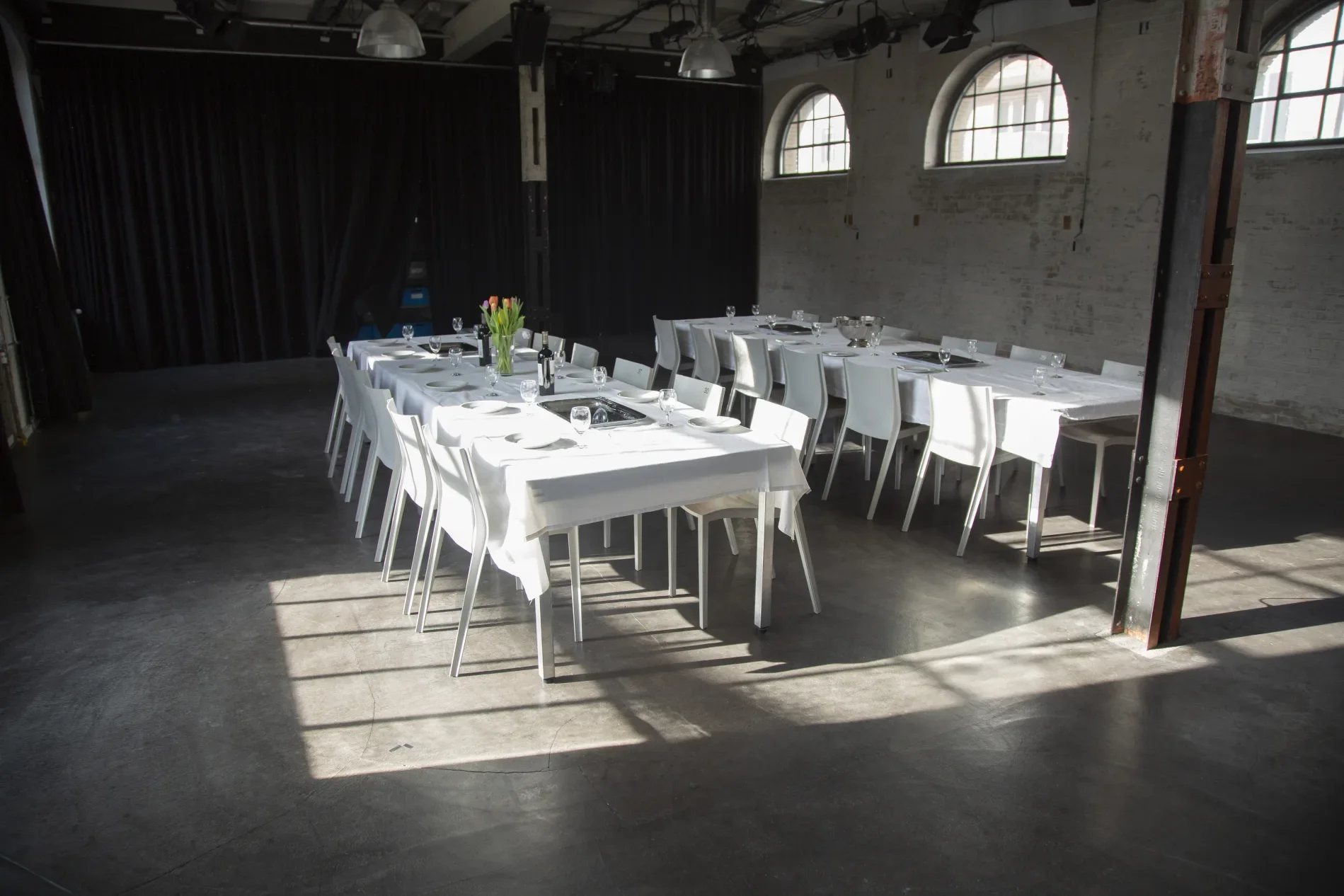 Ein festlich eingedeckter Tische im industrial Stil, mit weißen Tischdecken, Gläsern, Geschirr und Blumen in der Mitte, in einem Raum mit großen Fenstern und hellem Licht.