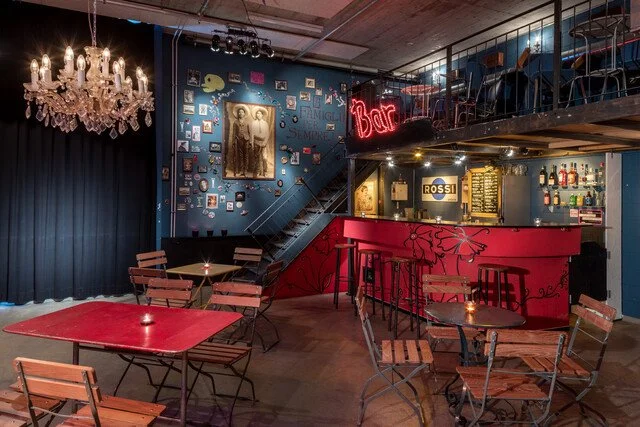 Innenansicht einer Bar mit Barhockern, Sitzbereichen, einer Treppe, einer Wanddekoration und einem Kronleuchter