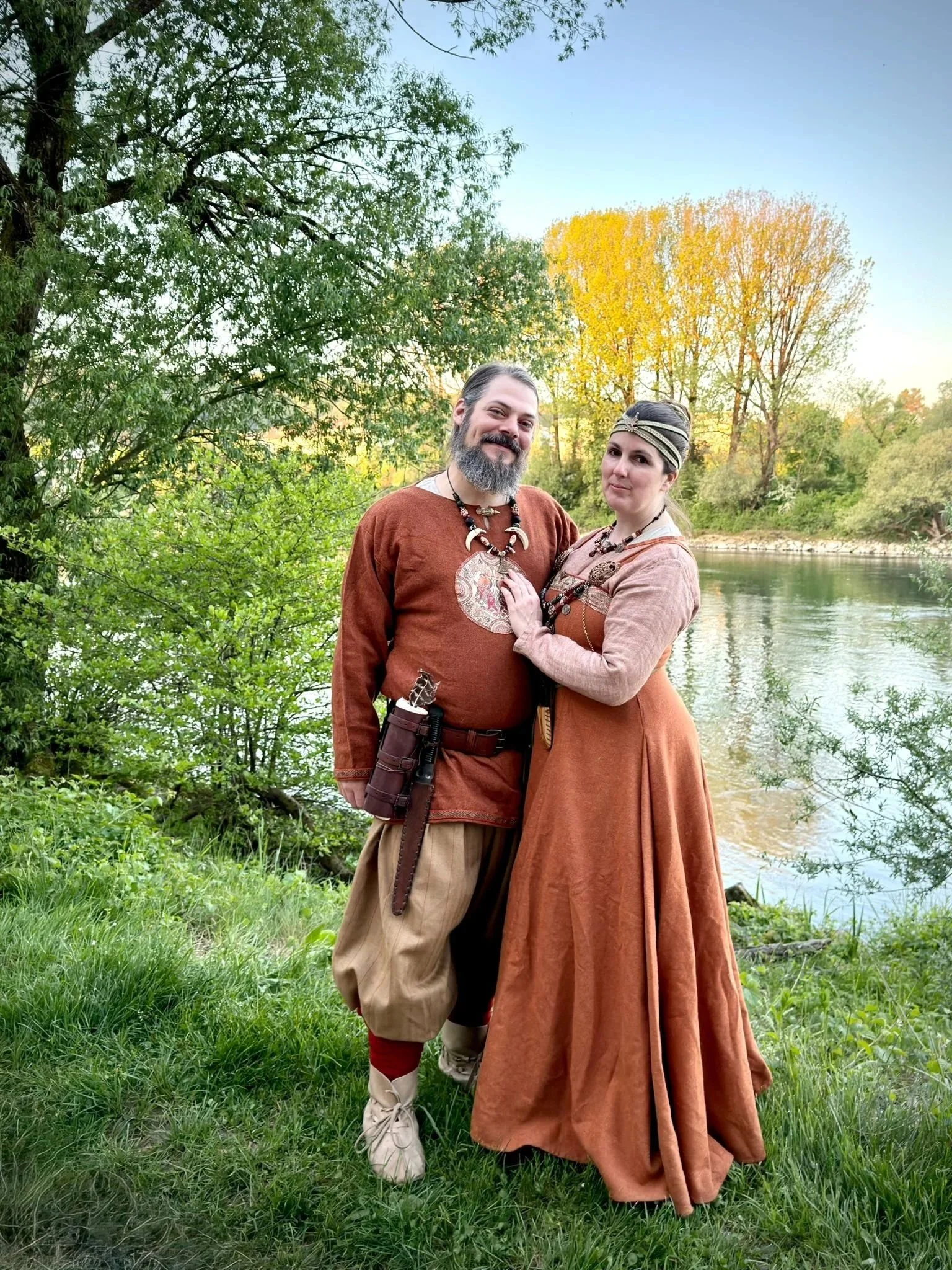 Ein Mann und eine Frau in mittelalterlicher Kleidung stehen am Flussufer in einer grünen, bewaldeten Umgebung, im Sonnenlicht bei Sonnenuntergang