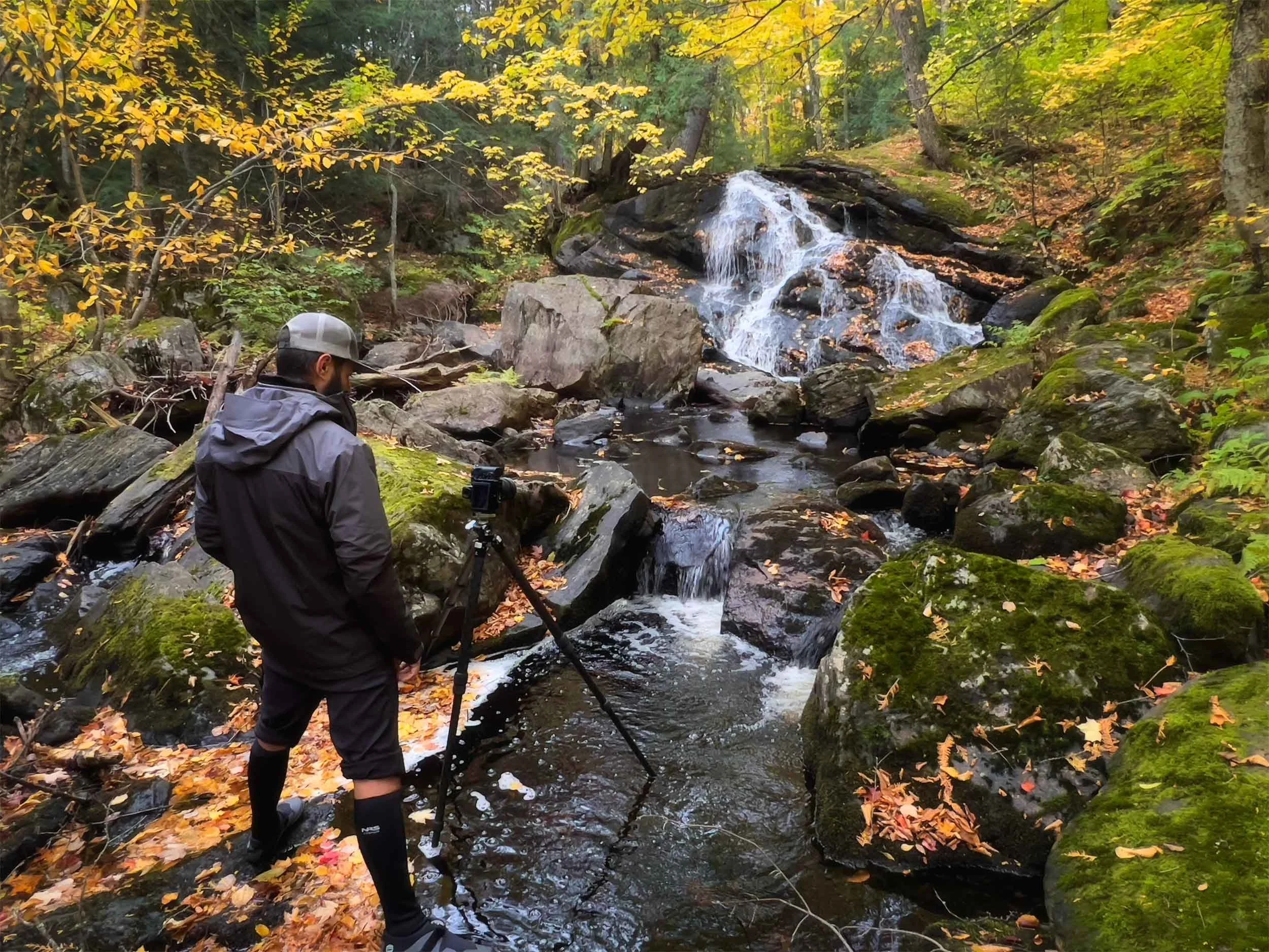 A person in outdoor clothing, including a gray cap and black waterproof boots, is filming or photographing a small waterfall in a forest with autumn leaves. The scene features rocks, a stream, and trees with yellow and green leaves.