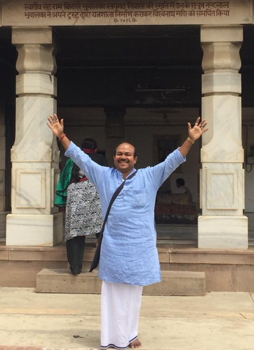 A man in traditional Indian attire with arms raised, smiling at a temple entrance with white marble columns and a Hindi inscription above.