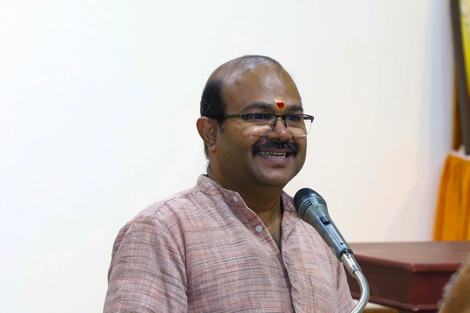 Man in glasses speaking at a microphone, wearing a traditional Indian kurta, with a small red and gold decorative mark on his forehead.