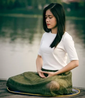 A young woman with long dark hair, wearing a white shirt, standing outdoors near a body of water, with a large green cloth or bag placed on a surface in front of her.