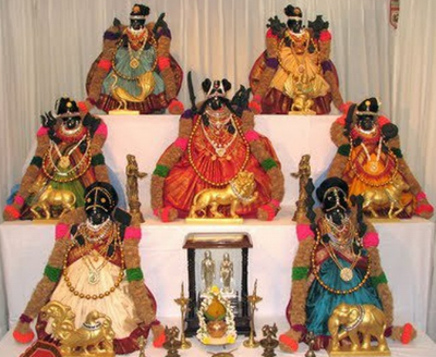 Collection of ornate Hindu deity statues dressed in colorful clothing, adorned with jewelry, and displayed on white platforms with a small table in front holding a framed picture and a Shiva Nataraja statue.