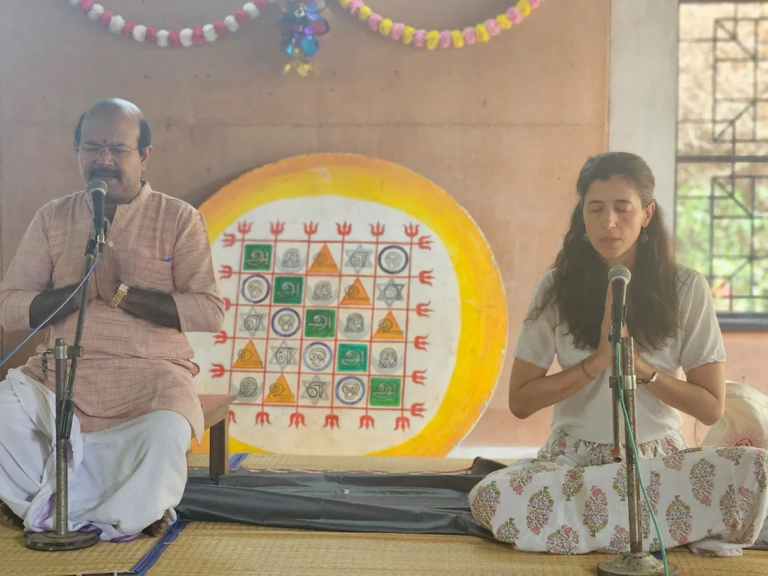 A man and woman sitting on a stage, both with microphones, engaged in prayer with closed eyes and hands folded. Behind them, there is artwork of a colorful geometric and symbolic design on a wall. The setting appears to be a traditional or spiritual 