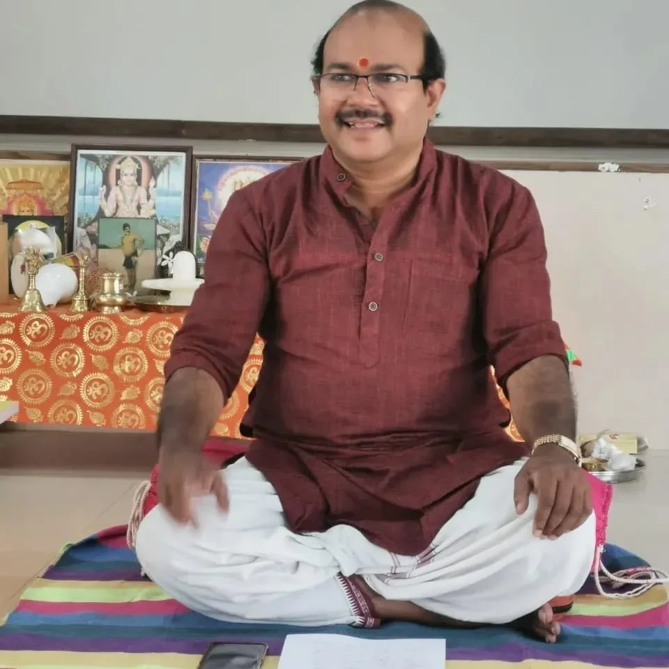 A man sitting cross-legged on a colorful mat, wearing glasses and traditional Indian attire with a red kurta and white dhoti, in front of a table with religious idols and pictures.