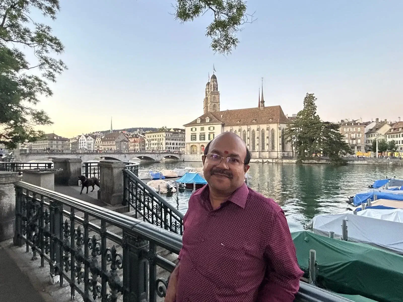 A man standing by a river with buildings and boats in the background, featuring a church with tall spires.