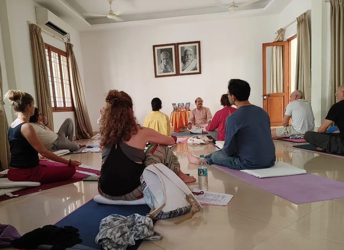 Group of people participating in a yoga or meditation session in a bright room with windows, framed pictures on the wall, and an instructor leading the session.