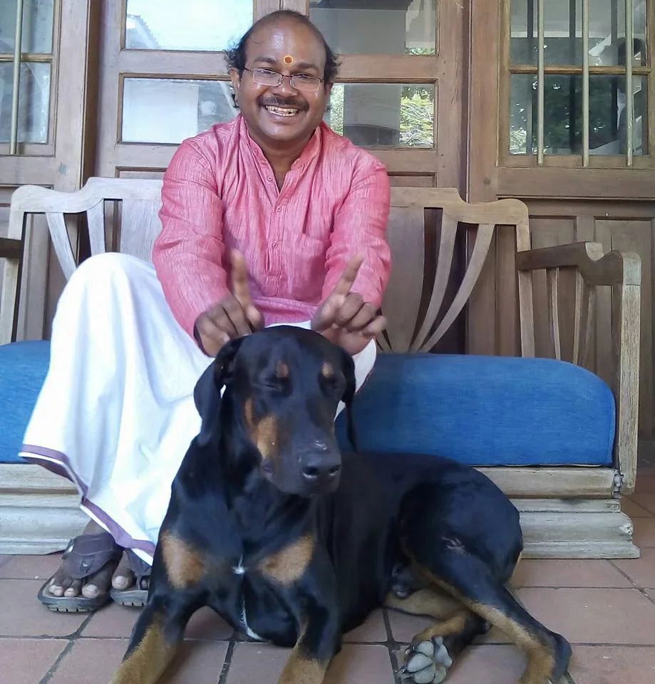 A man with glasses and a mustache, smiling and wearing traditional Indian clothing, sitting on a wooden bench with a black and tan dog lying in front of him on the floor.