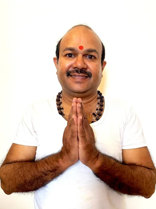 Man with a red dot on his forehead, wearing a white shirt, prayer beads around his neck, and clasped hands in a greeting pose.