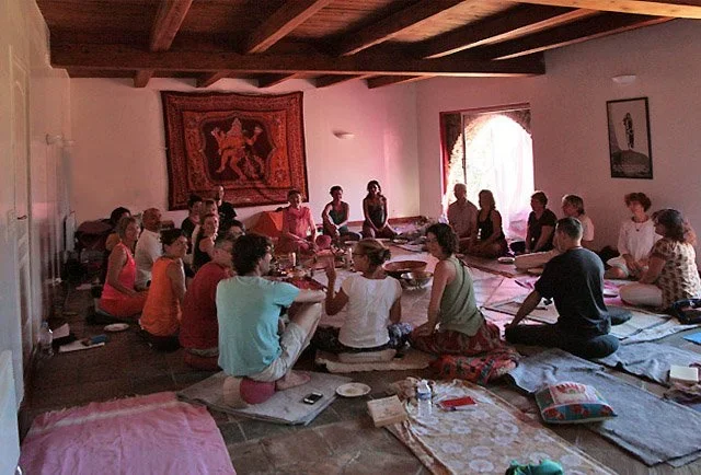 A group of people sitting cross-legged on mats in a large room, possibly engaging in a meditation or discussion session. The room has a wooden ceiling, a tapestry on the wall, and a window letting in natural light.