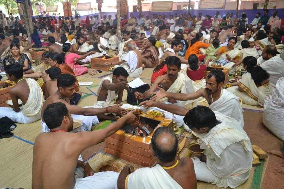 A large group of people participating in a traditional Hindu ceremony, sitting on the floor with offerings and religious items, some lighting a sacred fire, in an open-air space decorated with religious banners.