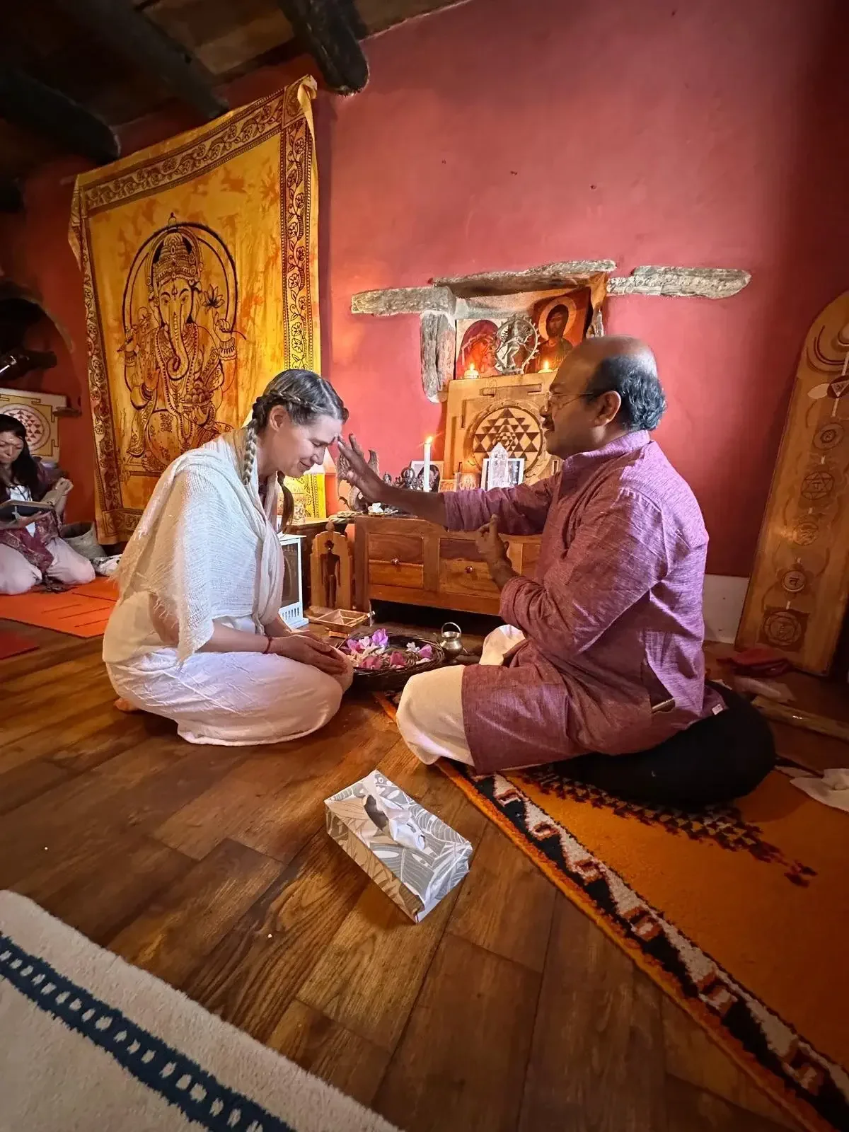 A woman dressed in white sitting on a carpet with her hands on her lap, receiving a blessing from a man in traditional clothing. They are in a room with religious icons, a lit candle, and decorative fabrics.