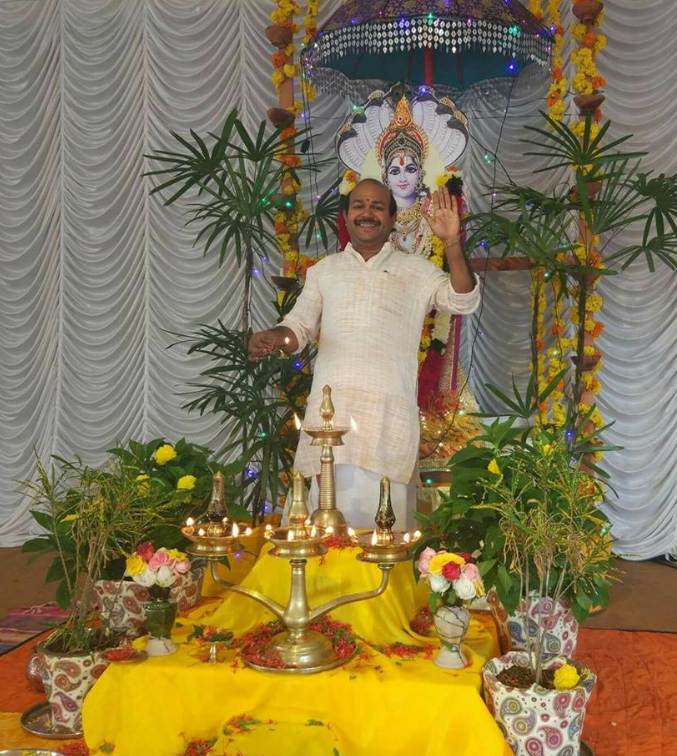A man standing on a decorated stage, smiling and waving in front of a Hindu deity idol, surrounded by potted plants and flowers, during a religious or festive celebration.