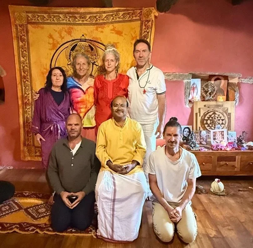 Group of seven people posing indoors with spiritual and decorative items, including a tapestry with a spiritual symbol, photographs, and statues in the background.