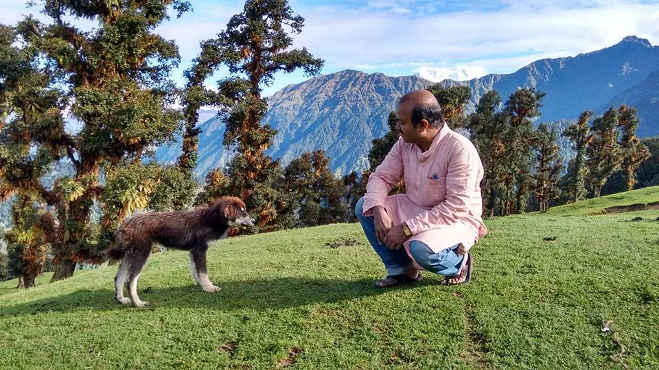 A man crouching down and smiling at a dog on a grassy hill with trees and mountains in the background.