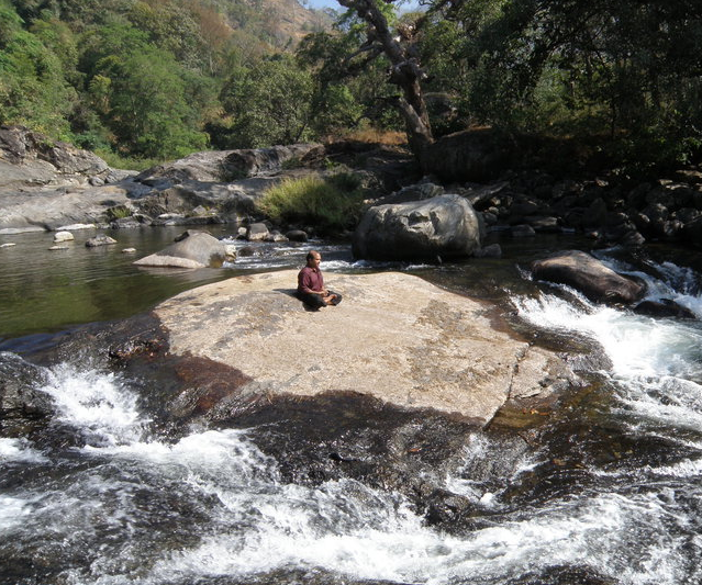 Person sitting meditating on a large flat rock in a flowing river surrounded by trees and rocks.