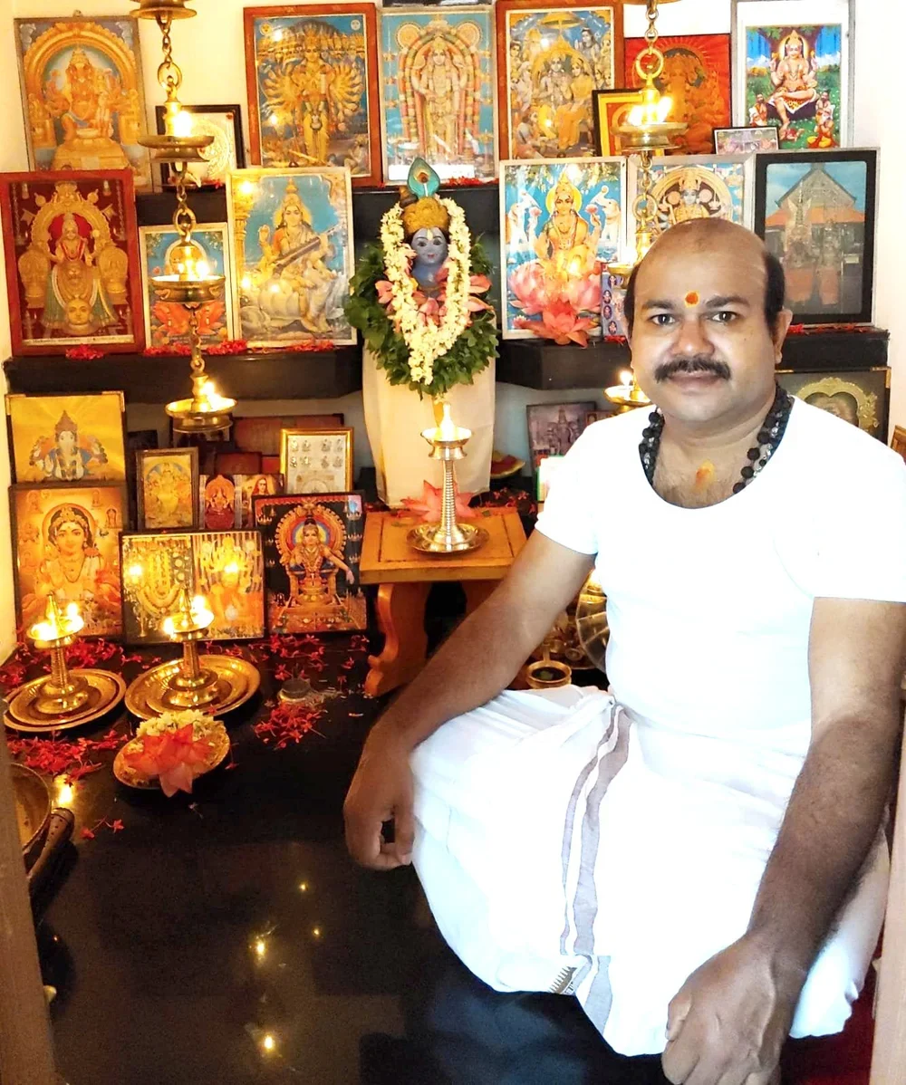 A man in traditional Indian attire participating in a Hindu prayer ceremony with colorful images and icons of Hindu gods and goddesses, lit lamps, and flower decorations in a room.