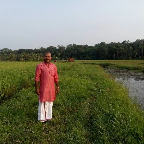 A man standing on a grassy path beside a body of water in a rural area with trees in the background, dressed in traditional Indian attire, smiling at the camera.