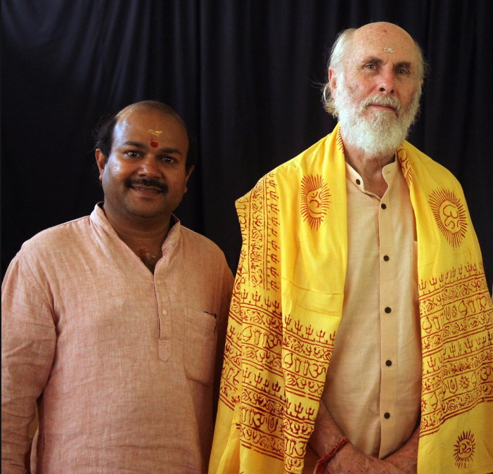 Two men standing side by side in front of a black background, one wearing a traditional outfit and a shawl with religious symbols, both with tilaks on their foreheads.