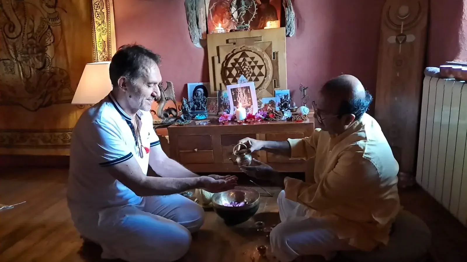 Two men performing a traditional ritual in a decorated room, with religious and spiritual items displayed on a table behind them.