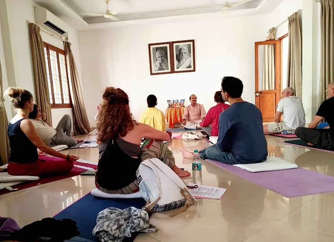 A group of people participating in a meditation or yoga session in a bright, spacious room with white walls, wooden window frames, and a door. They are sitting on yoga mats or cushions, facing a man at the front who appears to be instructing the session. Two framed portraits are hanging on the wall behind him.