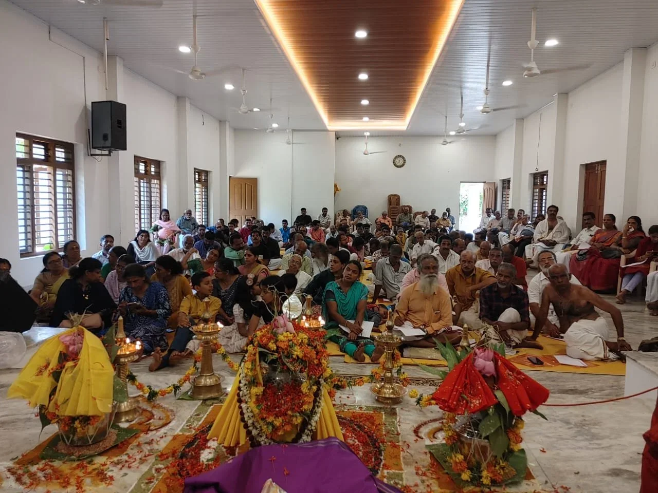 A large gathering of people participating in a religious or cultural ceremony inside a spacious hall, with decorations, flower arrangements, and offerings at the front.