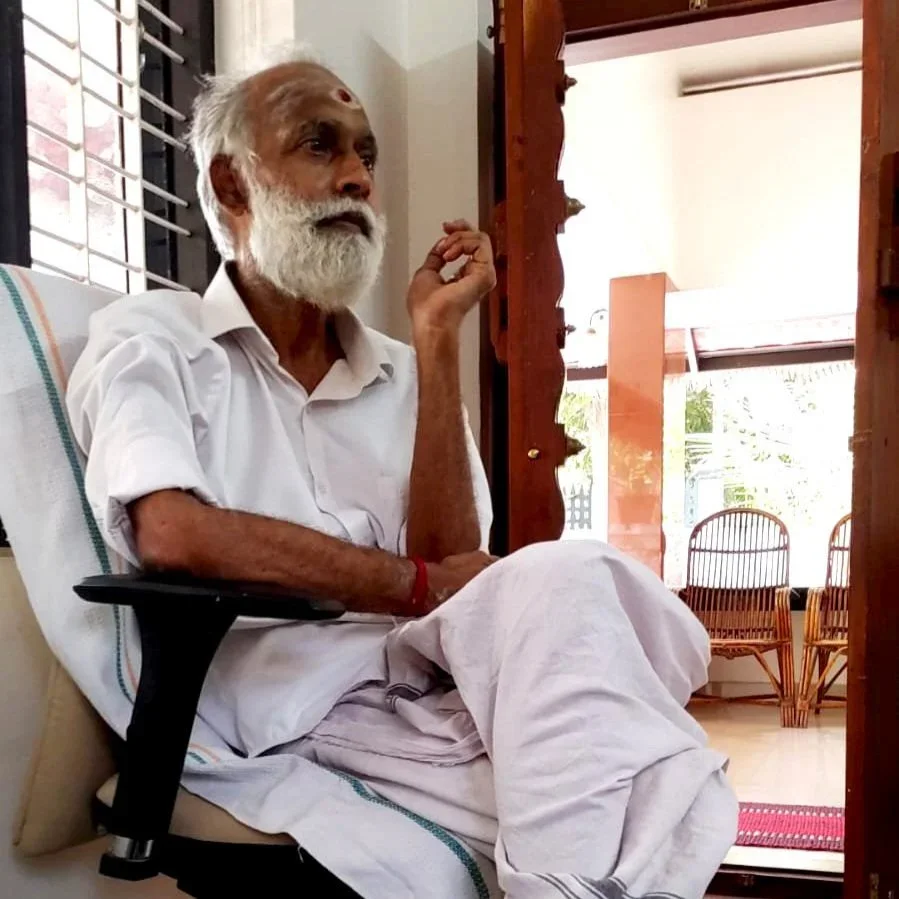 An elderly man with white hair and beard sitting on a chair in a room, wearing traditional Indian attire, looking to the side with a calm expression.
