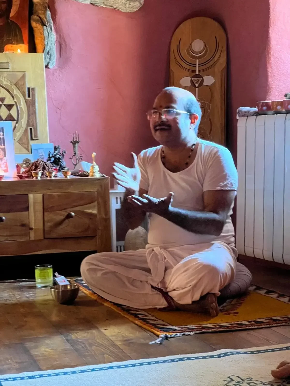 A man sitting cross-legged on the floor, engaged in a spiritual or religious ceremony, with prayer beads around his neck, gesturing with his hands, and surrounded by religious icons and items on a wooden altar against a pink wall.