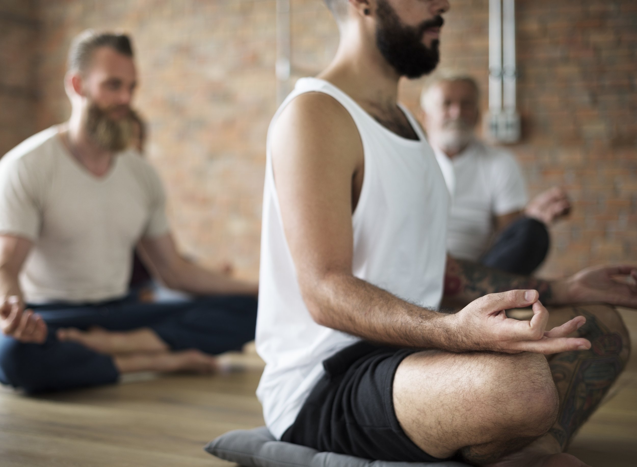 A group of men practicing meditation in a yoga studio with brick walls. They are seated cross-legged on mats, with eyes closed, focusing on their breathing.