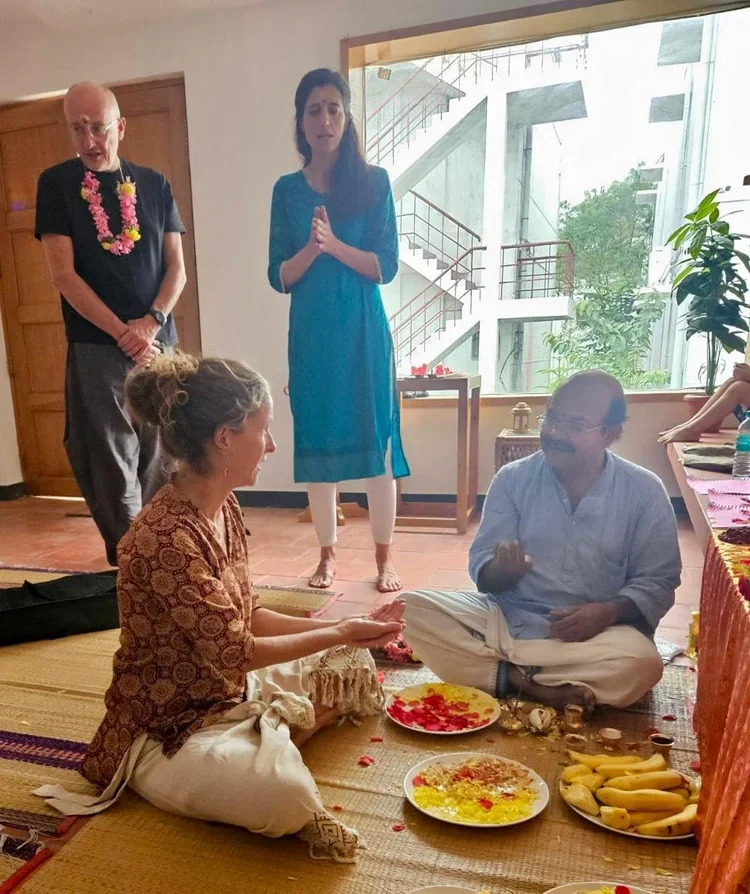 A woman and a man sitting on a prayer mat engaged in a ritual, with various food offerings and flower petals in front of them. Two people stand behind them, one a woman in a blue dress with hands pressed together in prayer, and the other a man with a